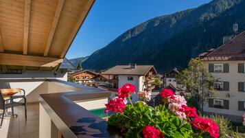 Ein Balkon mit Holzboden, einem Tisch und Stühlen. Es stehen Kübel mit roten Blütenpflanzen darauf. Im Hintergrund sind Berge und Gebäude zu sehen.