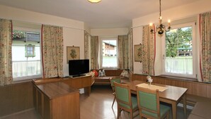 A cozy living room with a wooden bench, a dining table, and chairs. There is a television on a wooden cabinet, and the room has large windows with curtains. - Apartment Rauter by Interhome (Mayrhofen)