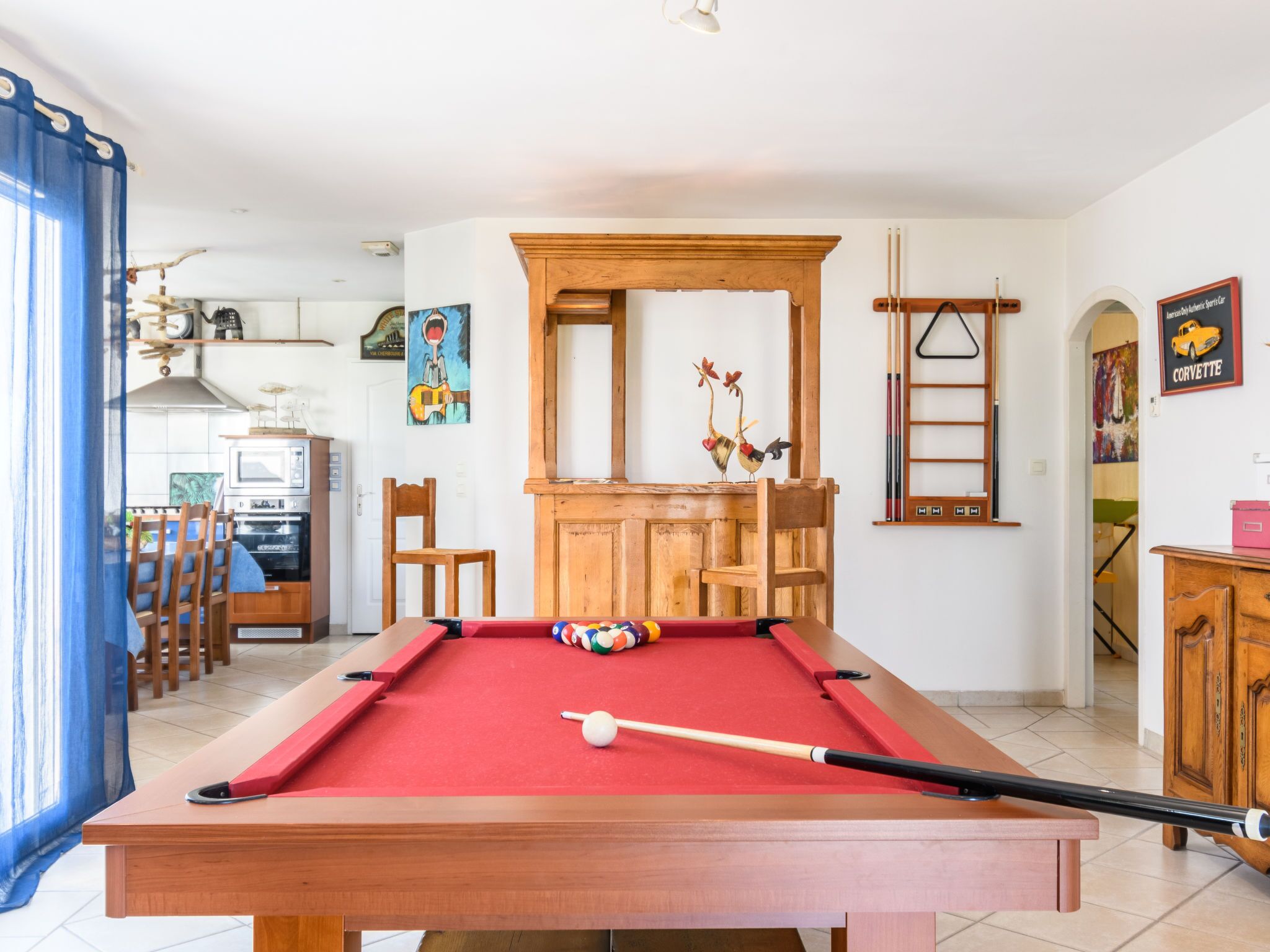 Une table de billard dans une pièce où l’on aperçoit un buffet en bois, des chaises et, au fond, un coin repas.