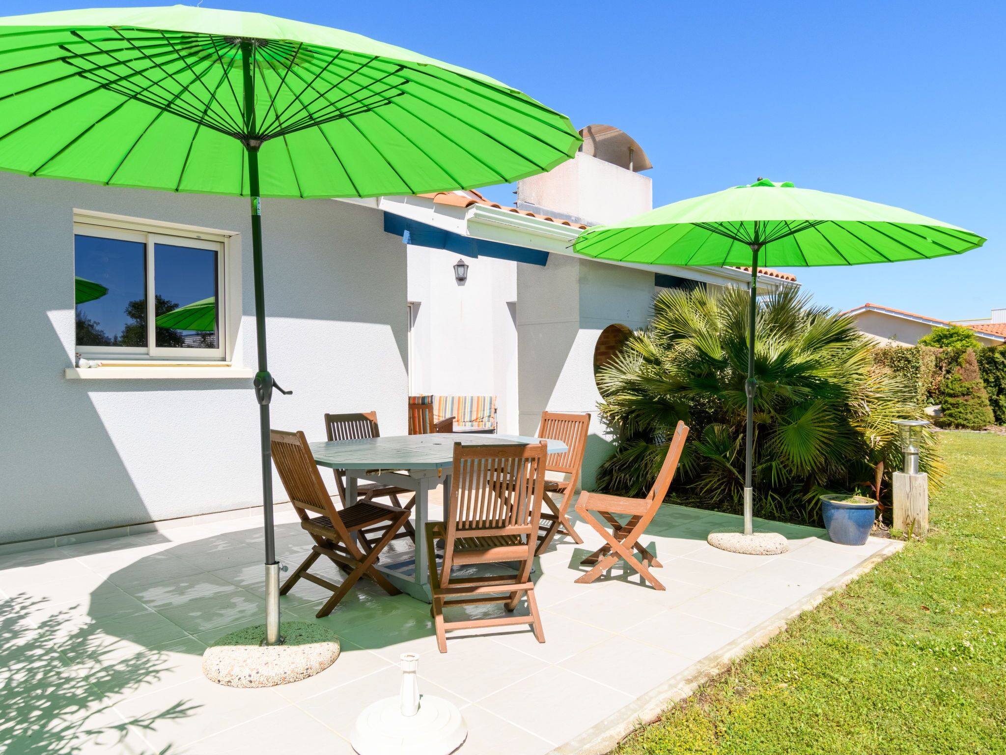 An outdoor patio with a table and chairs, two green umbrellas, and a white building with a window.