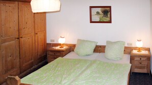 Un lit en bois recouvert d’une couvre-lit à motifs floraux verts, deux tables de chevet avec des lampes, une armoire en bois et, au mur, un tableau représentant un paysage.