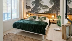 Un dormitorio con cama, mesita de noche, lámpara, cuadro de árboles y ventana con persianas.