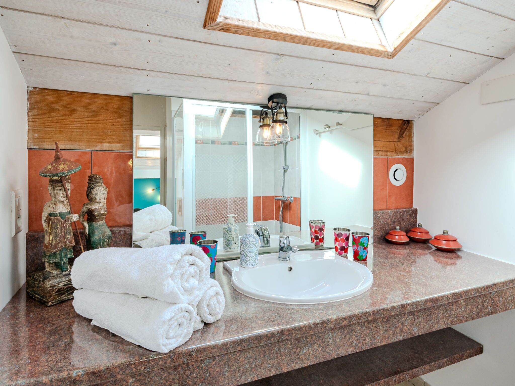 A bathroom with a large mirror, a sink, and a shower. There are towels stacked on the counter, and decorative items like a Buddha statue and a small Buddha figure.
