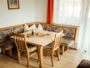 Un coin repas comprenant une table en bois, quatre chaises, un banc avec des coussins à motifs et une fenêtre avec des rideaux.