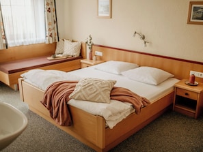 A neatly made bed with white linens and a brown throw blanket, a wooden headboard, a bedside table with a lamp, and a window with curtains.