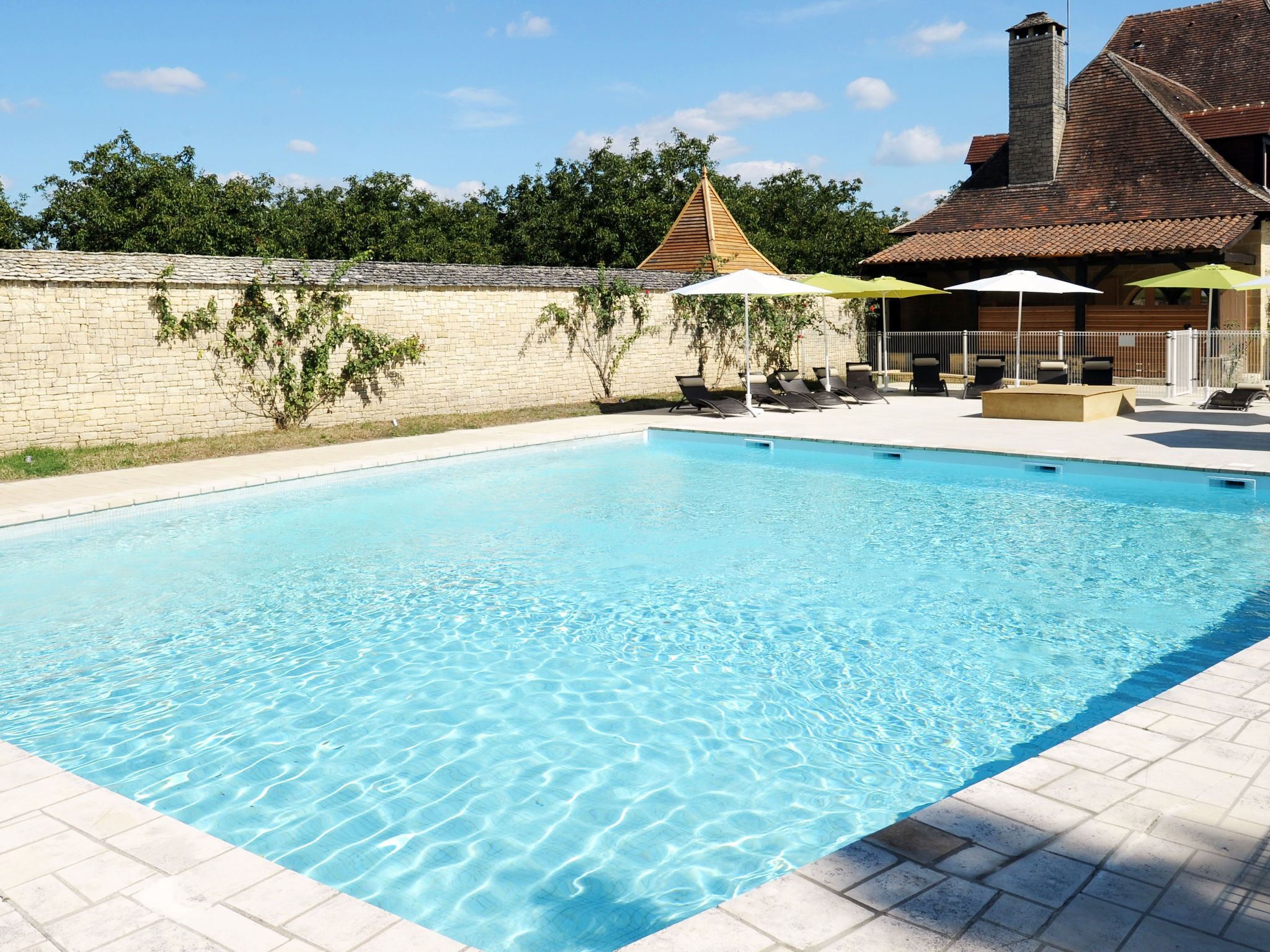 A rectangular swimming pool with clear blue water, surrounded by a tiled deck. There are lounge chairs and umbrellas, and a brick wall with greenery in the background.