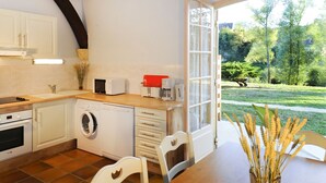 A kitchen with a washing machine, microwave, and coffee maker, a dining table, and a view of a garden.