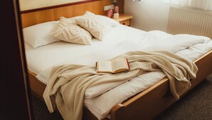 A neatly made bed with a book and a blanket, a bedside table with a vase, and a window with curtains.