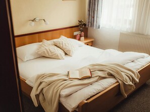 Ein ordentlich bezogenes Bett mit einem Buch und einer Decke, ein Nachttisch mit einer Vase und ein Fenster mit Vorhängen.
