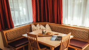 A corner seating area with a wooden table and chairs, a bench with cushions, and a window with red curtains.