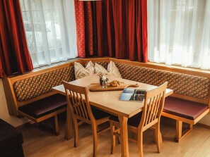 Eine Sitzecke mit Holztisch und Stühlen, eine Bank mit Kissen sowie ein Fenster mit roten Vorhängen.