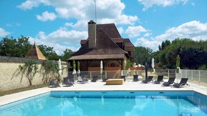 A swimming pool with lounge chairs and umbrellas, surrounded by a stone wall and a wooden building with a tiled roof.