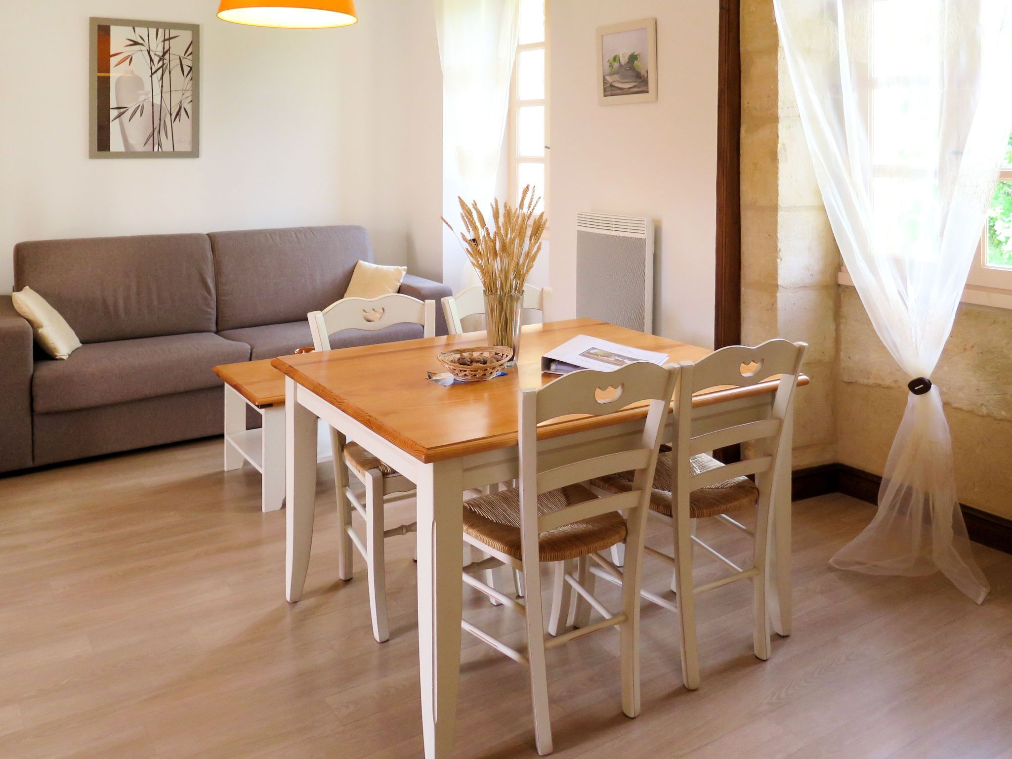 Uma área de refeições com uma mesa de madeira e cadeiras brancas, um sofá cinzento e um radiador na parede.