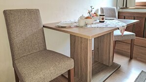 Un coin repas avec une table en bois et deux chaises rembourrées. Une nappe à motifs floraux recouvre la table, et on y trouve divers objets, notamment une théière blanche, une tasse, un bol contenant du pain et des fruits, et un pot de confiture.