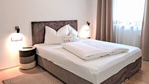 Un lit bien fait, avec du linge de lit blanc et des coussins décoratifs, une lampe de chevet et une table de chevet ronde à motifs rayés.