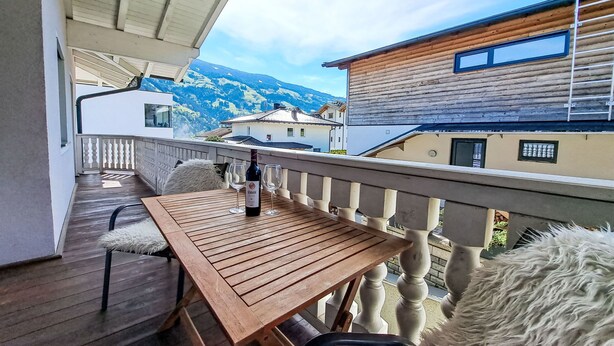 Una mesa de madera con una botella de vino y dos copas en un balcón con vista a una montaña y edificios.