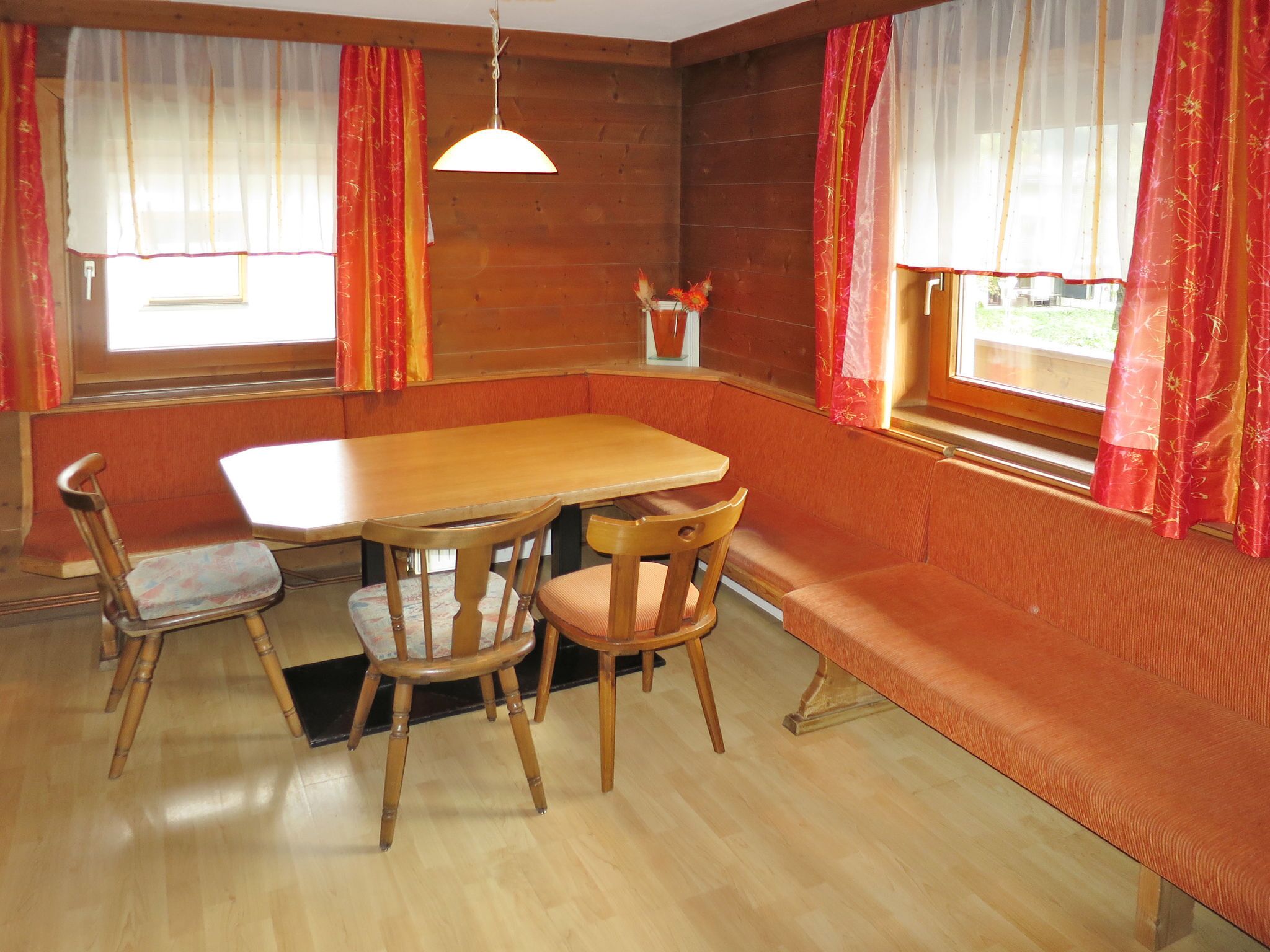 Un coin repas avec une table en bois et quatre chaises, un banc et deux fenêtres ornées de rideaux rouges.