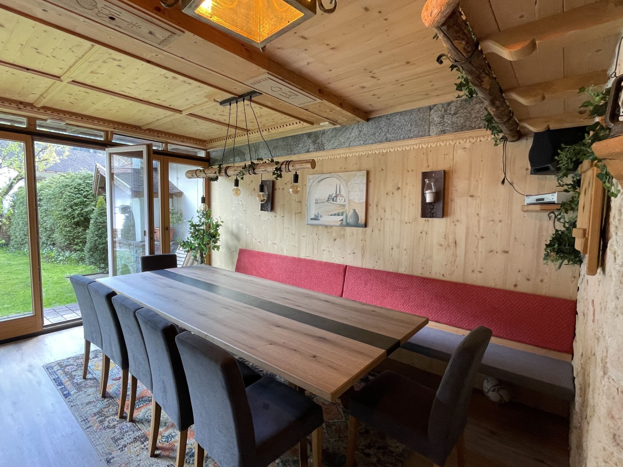 Un espace repas avec une longue table en bois, des chaises noires et un banc rouge. Des luminaires suspendus et un tableau ornent le mur.