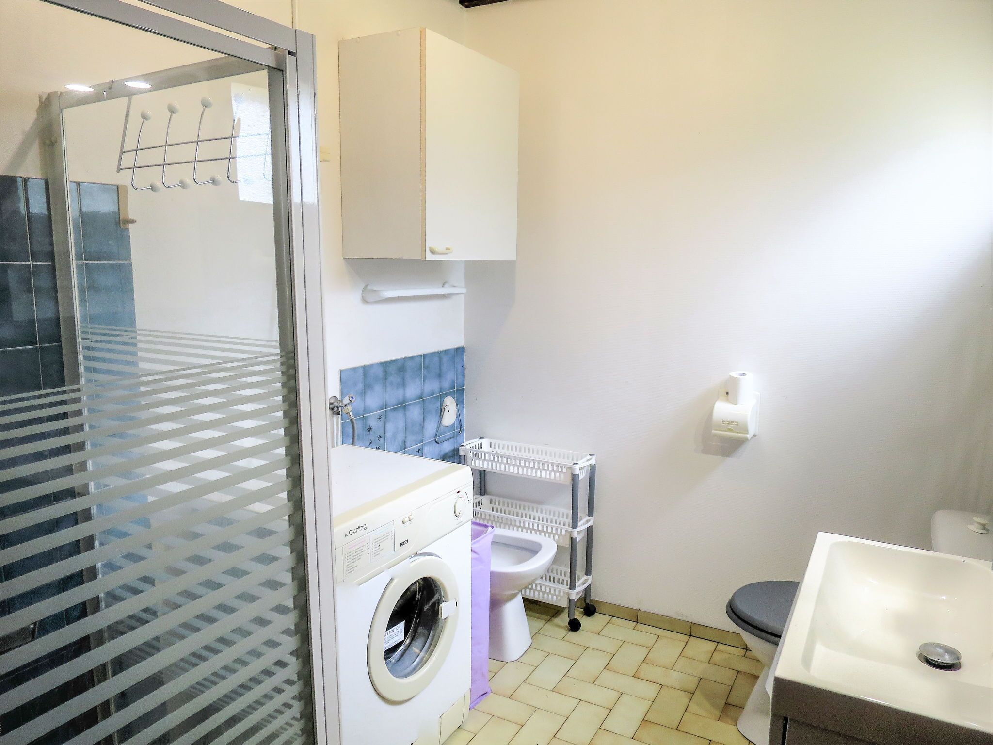 A compact hotel bathroom with a washing machine, a toilet, a sink, and a small window with blinds.