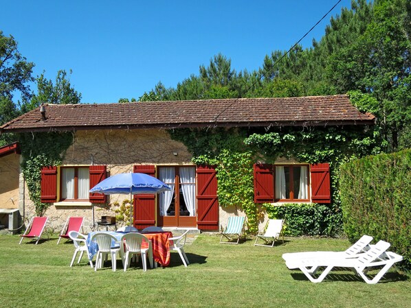 A rustic house with a tiled roof, red shutters, and a garden with lawn chairs and a table.