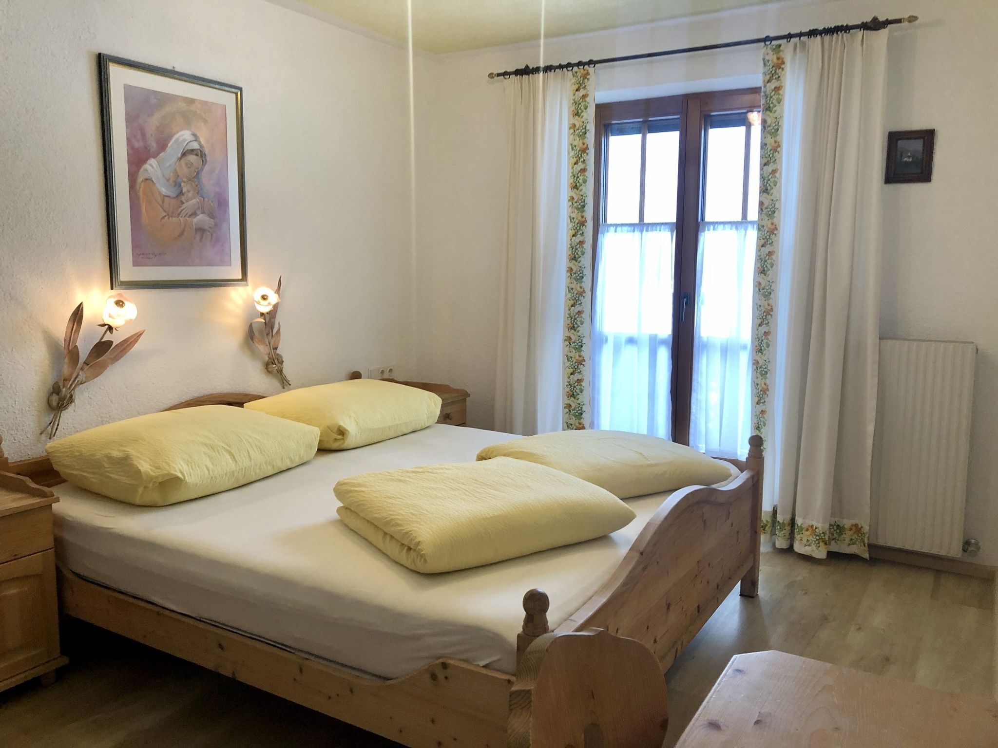 Un dormitorio con cama de madera, dos mesitas de noche, una mesita auxiliar, un cuadro en la pared y una ventana con cortinas florales.