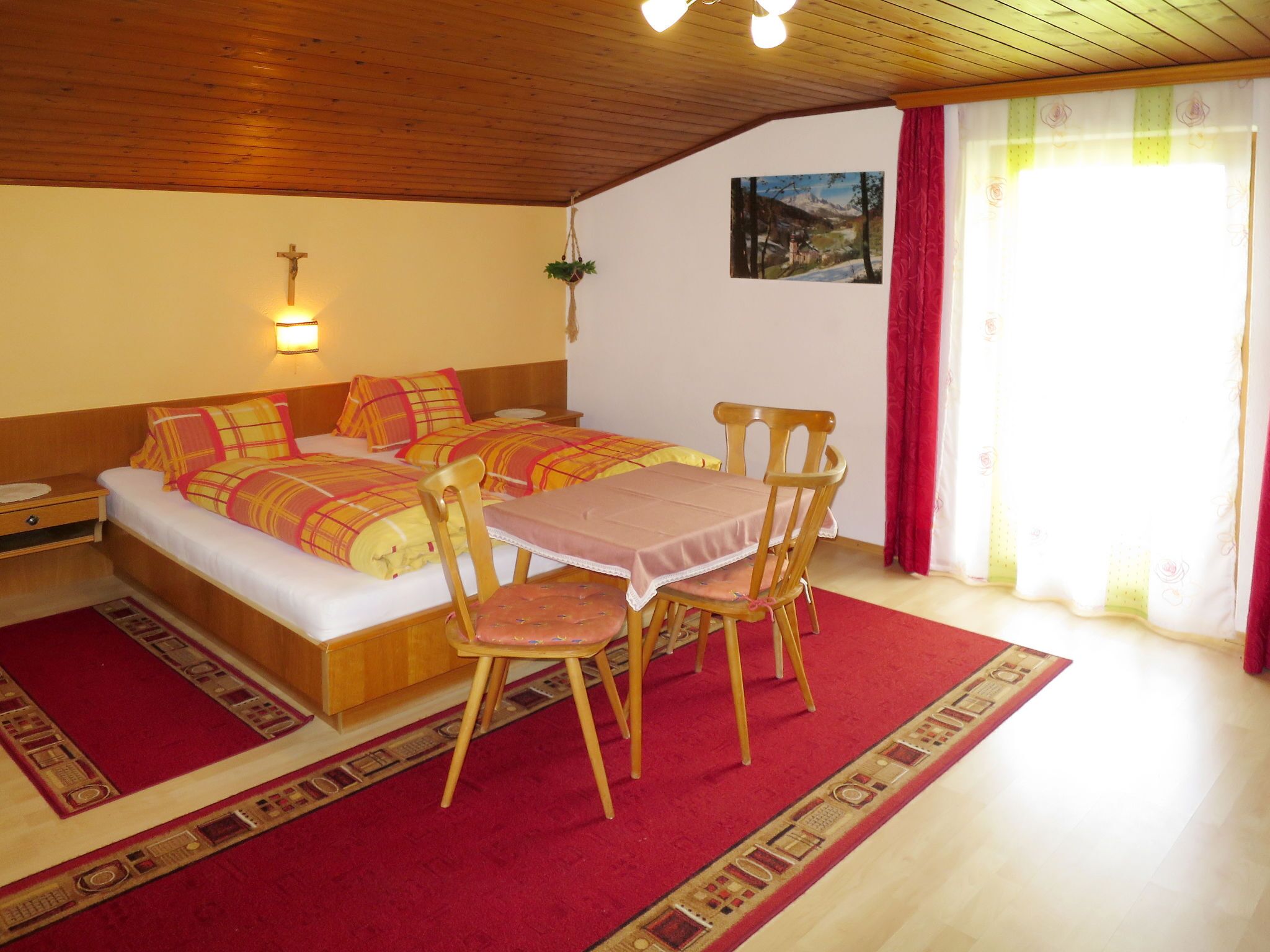 A cozy room with a bed, a table with chairs, a cross on the wall, and a painting.