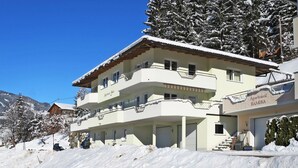 Um edifício com várias andares e varandas, com a placa "Apartment Ramona", rodeado por árvores cobertas de neve e um céu azul límpido.