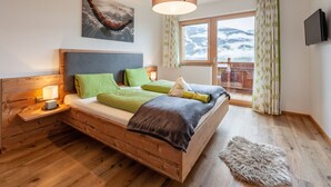 Una cama de madera con ropa de cama verde y gris, un televisor montado y una ventana con vistas a las montañas.