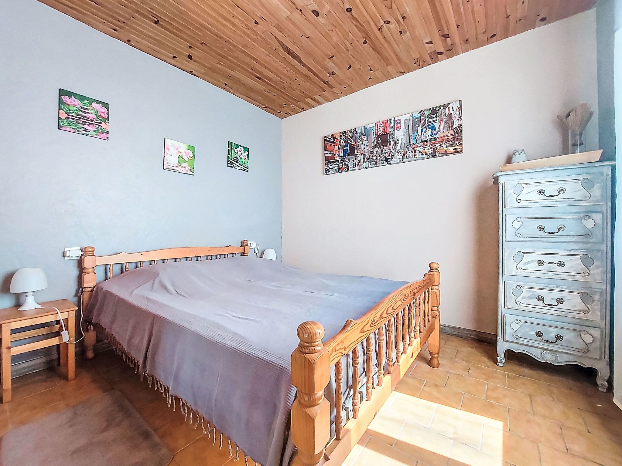A bedroom with a wooden bed, a nightstand with a lamp, a dresser with drawers, and a large cityscape painting on the wall.