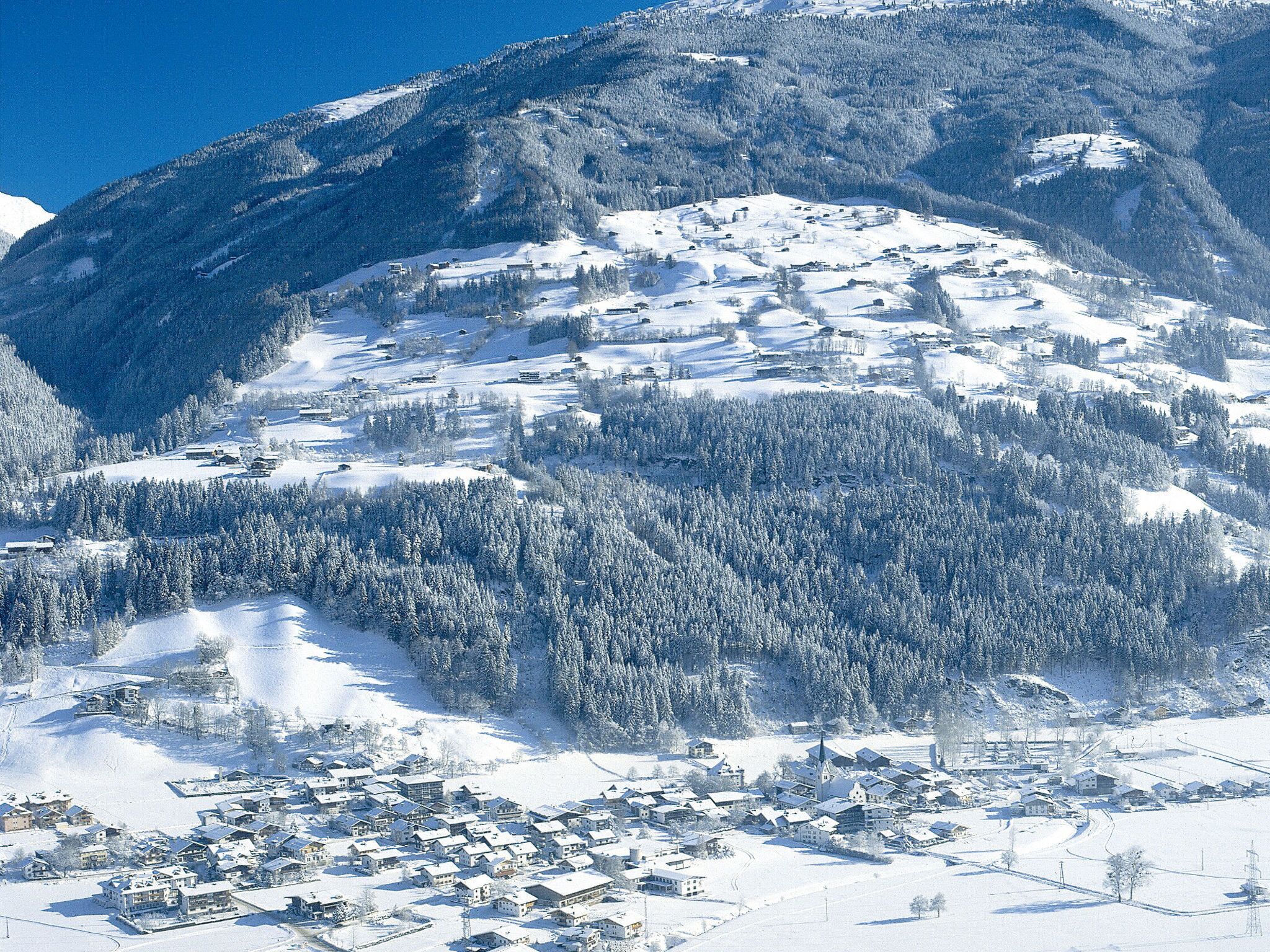 A snow-covered mountain village with buildings clustered at the base, surrounded by dense forests and steep slopes.