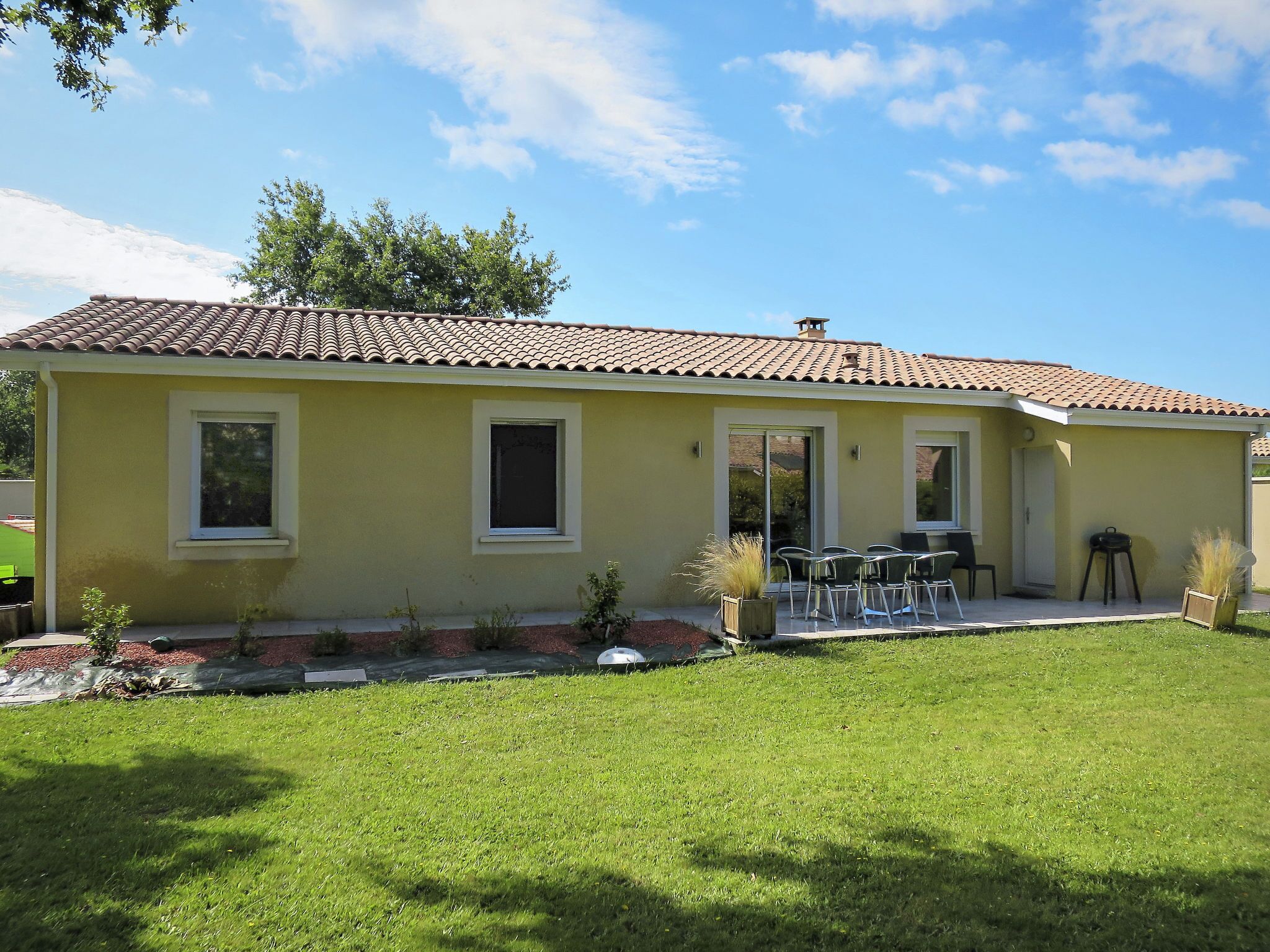 Uma casa amarela com telhado de telhas, um pátio com cadeiras e um pequeno jardim.