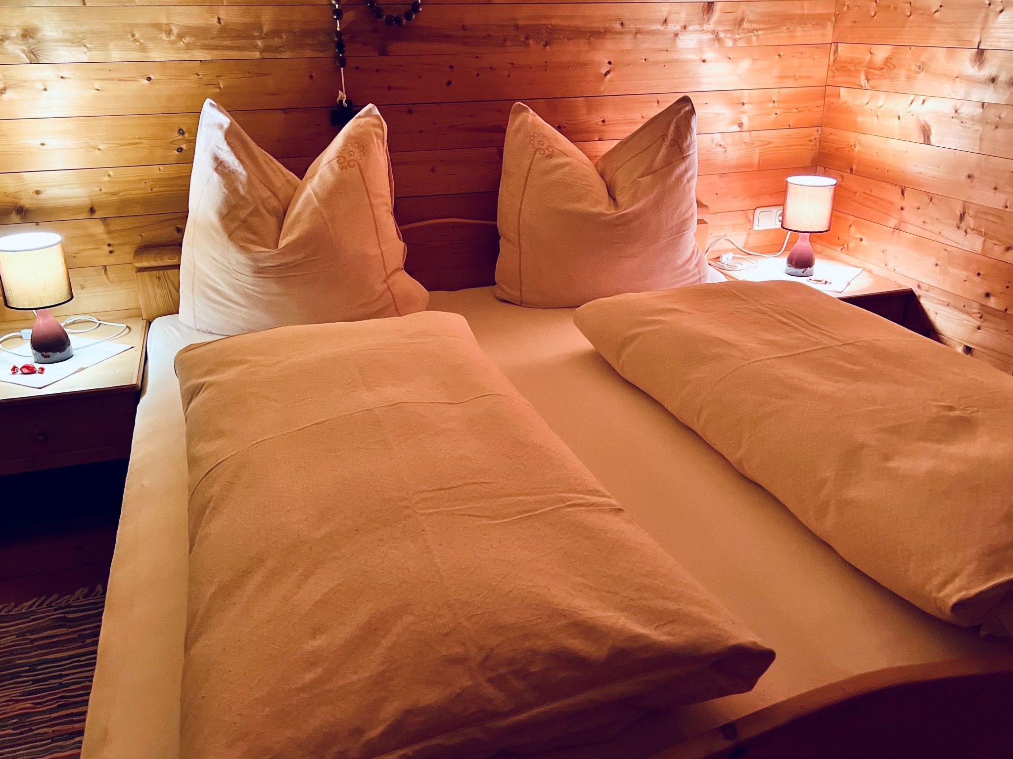 Ein Zimmer einer Holzhütte mit einem Bett, zwei Kissen, einem Nachttisch mit Lampe und einer Wandleuchte.