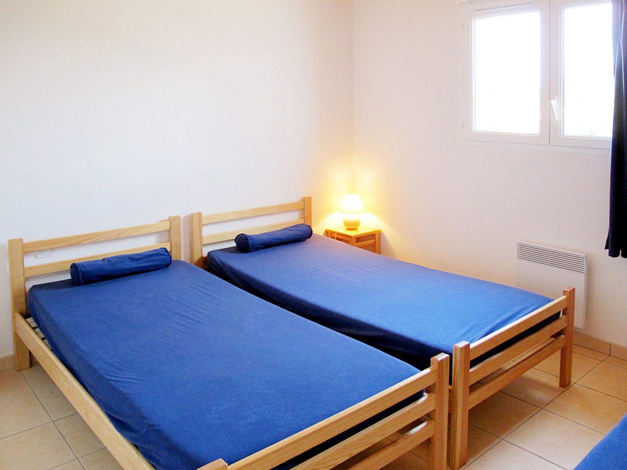 Una stanza piccola e pulita con due letti di legno, un comodino con una lampada e una finestra con tende.