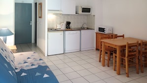 A compact kitchen with white appliances, a dining table with chairs, and a blue patterned sofa. - Les Hameaux de l'Océan 22 by Interhome (Carcans Plage)