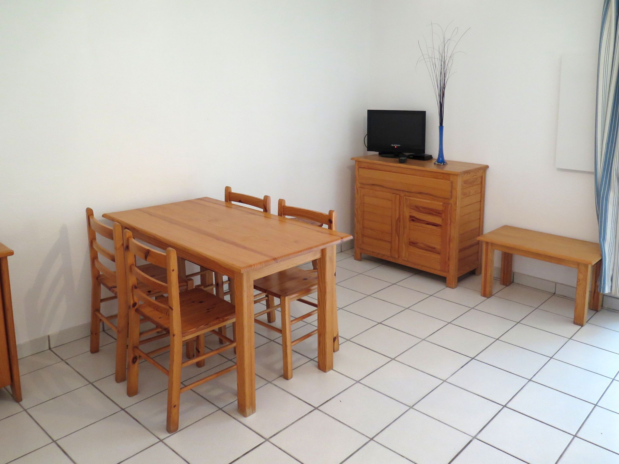 Ein Zimmer mit einem Holzesstisch, vier Stühlen, einer kleinen Holzkommode mit einem Fernseher und einer Bank.