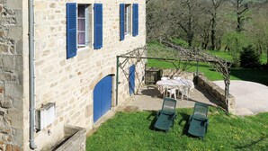 A stone building with blue shutters, a patio with a table and chairs, and a garden with a pergola.