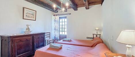 A room with two beds, a wooden dresser, a chandelier, a window with curtains, and a lamp on a side table.