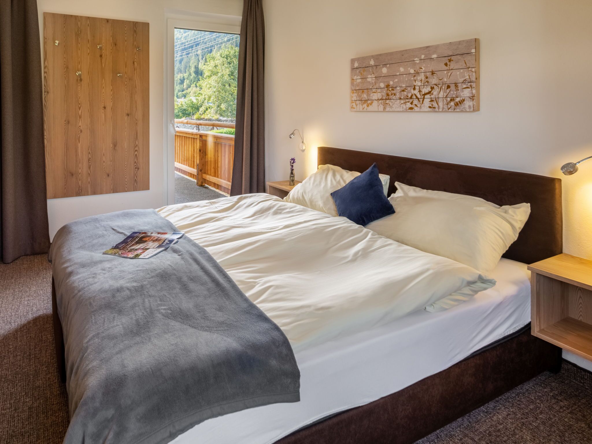 Ein ordentlich bezogenes Bett mit weißer und grauer Bettwäsche, ein Holzkopfschild und ein Nachttisch mit Lampe. Eine Holztür und ein Fenster mit Blick auf Grünpflanzen sind zu sehen.