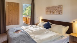 Un lit bien fait, avec une literie blanche et grise, une tête de lit en bois et une table de chevet avec une lampe. On aperçoit une porte en bois et une fenêtre donnant sur un paysage verdoyant.