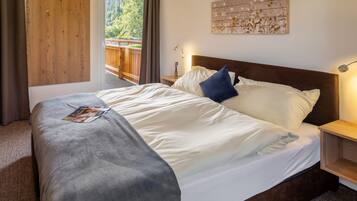 Ein ordentlich bezogenes Bett mit weißer und grauer Bettwäsche, ein Holzkopfschild und ein Nachttisch mit Lampe. Eine Holztür und ein Fenster mit Blick auf Grünpflanzen sind zu sehen.