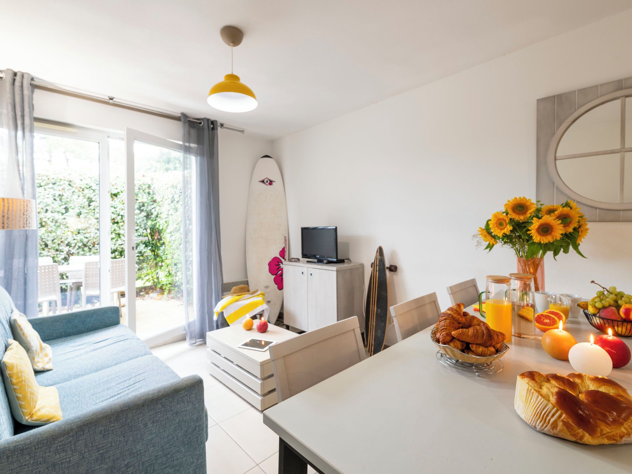 Un salon lumineux avec un canapé, une table à manger dressée pour le petit-déjeuner, une planche de surf et un téléviseur posé sur un meuble.