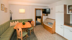 Un espace de vie compact comprenant une table à manger, deux chaises, un canapé, un téléviseur fixé au mur et une kitchenette équipée d’un évier et de placards.