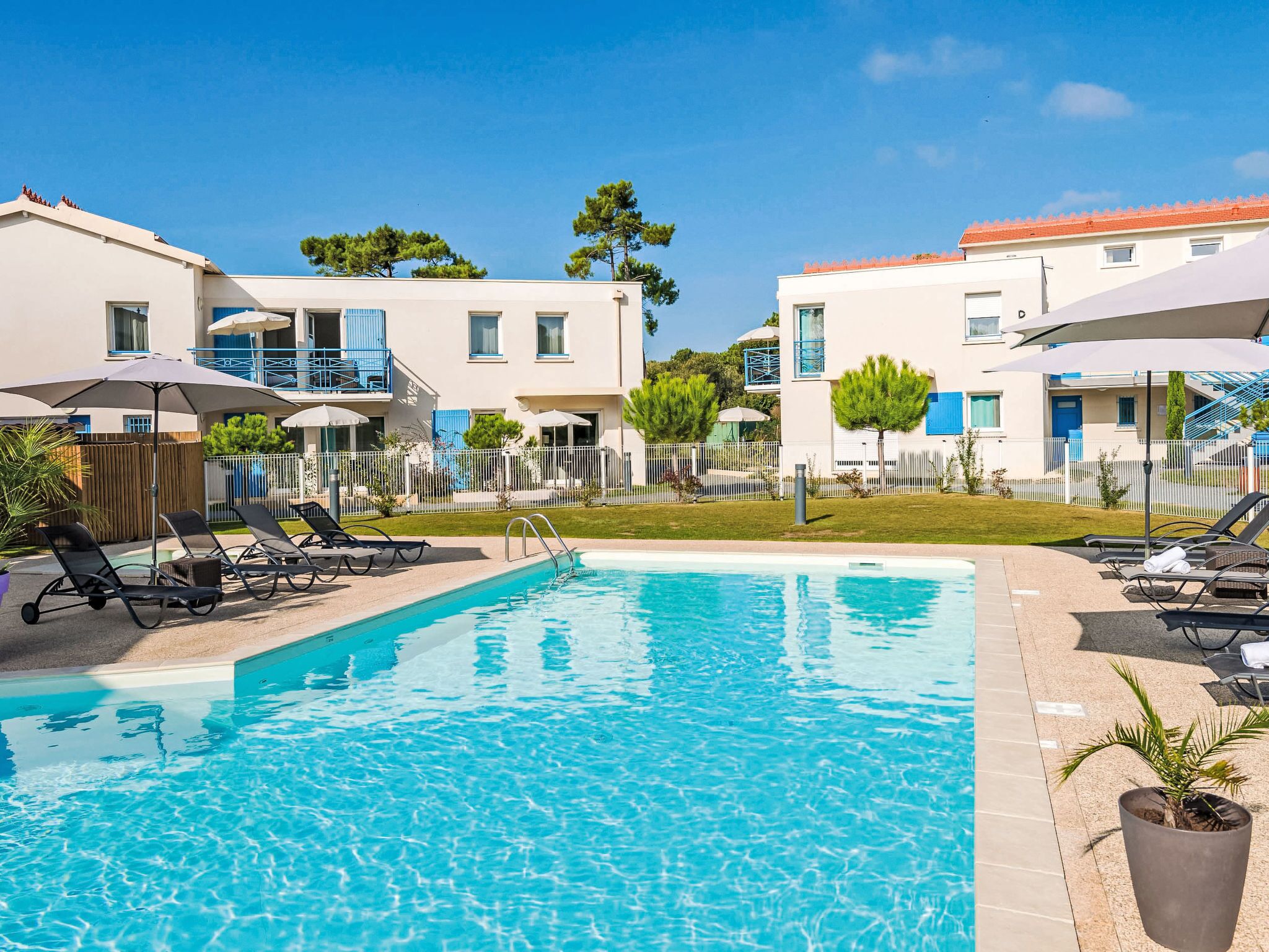 Una piscina con sillones de descanso y sombrillas, rodeada de edificios con balcones y un patio cercado.