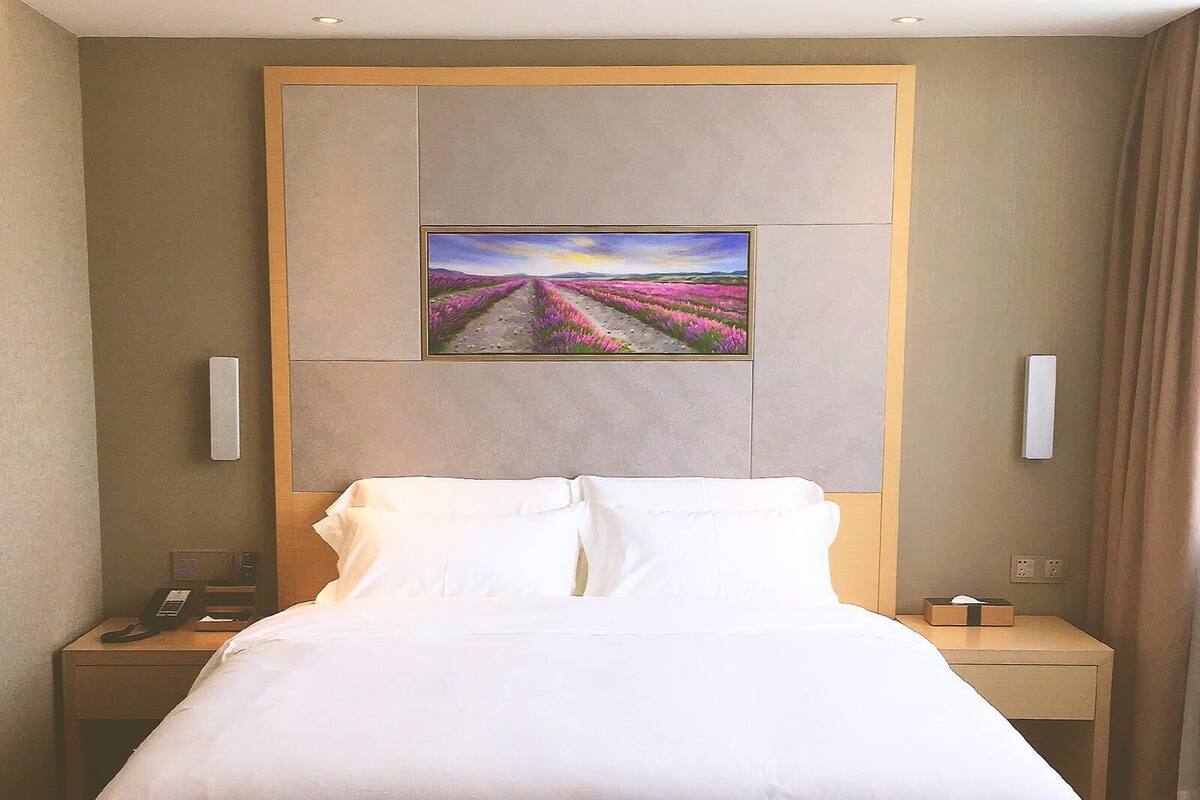 Una habitación de hotel con una cama grande, una mesita de noche con un teléfono, y un cuadro de un paisaje en la pared.