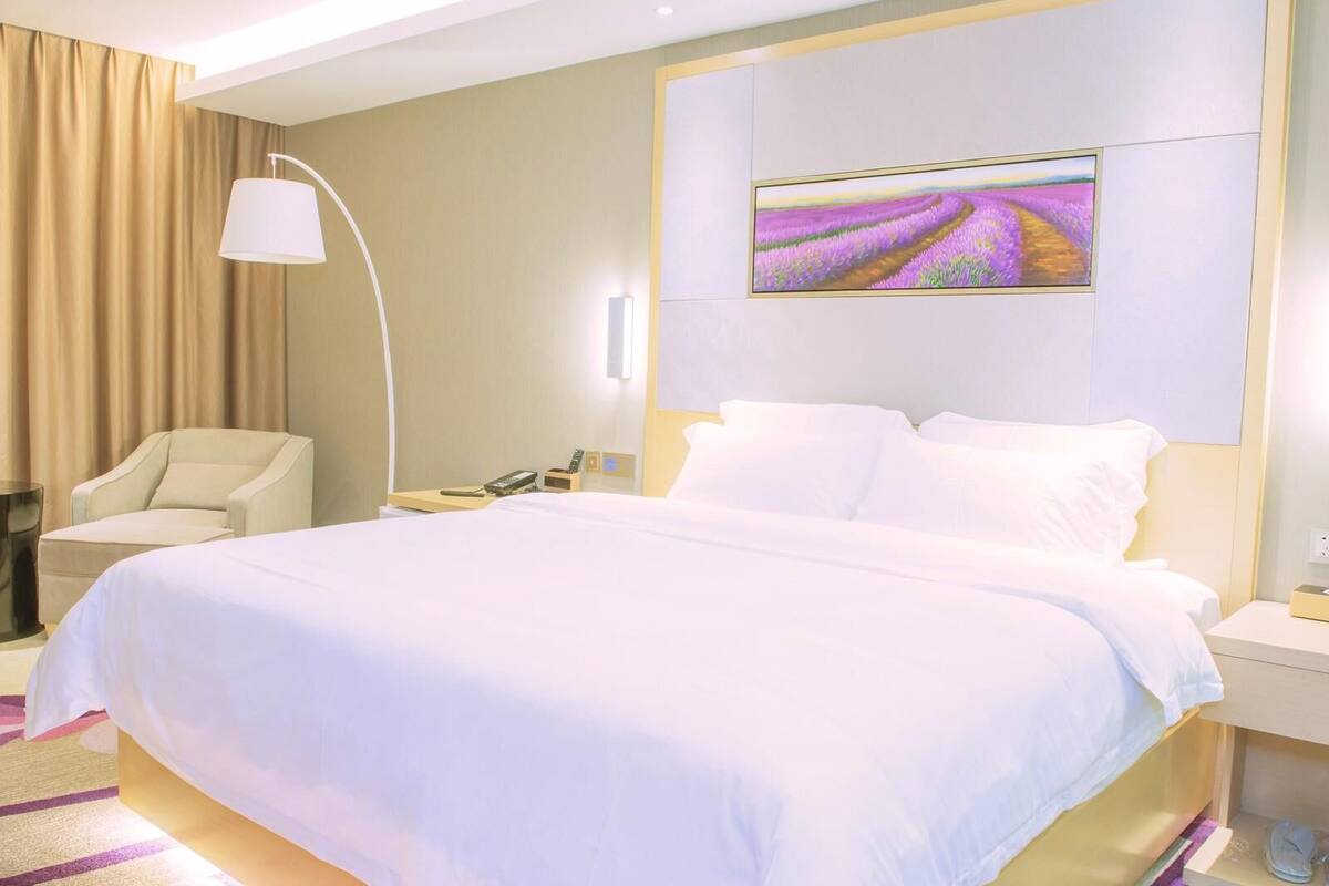Una habitación de hotel con una cama grande, una silla, una mesita de noche con lámpara y un cuadro en la pared.