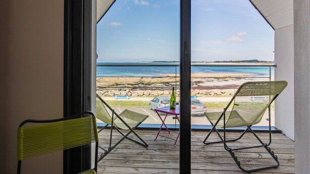 Varanda com duas cadeiras e uma pequena mesa, com vista para uma praia e um mar claro, além de uma linha de costa distante.