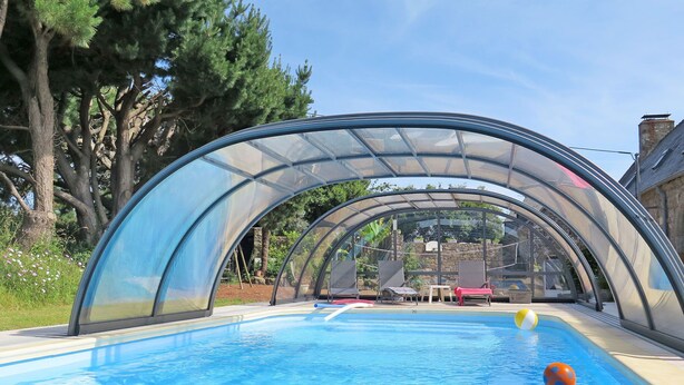 Una piscina con techo transparente, rodeada de árboles y un muro de piedra.