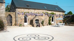 Un edificio de piedra con entrada de tejas, mesas al aire libre y flores moradas colgantes.