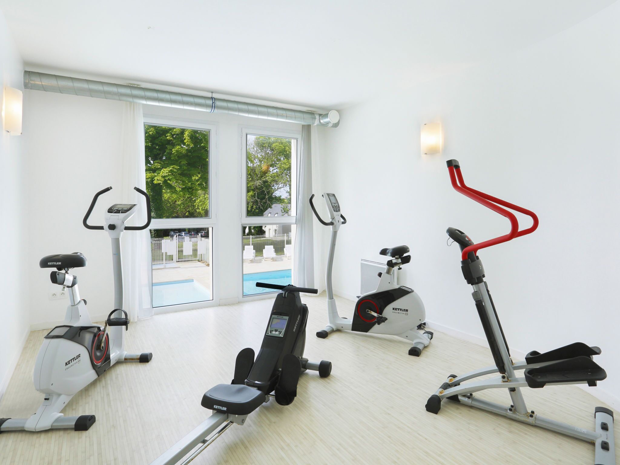 Ein Fitnessstudio mit verschiedenen Trainingsgeräten, großen Fenstern und Blick auf einen Swimmingpool.
