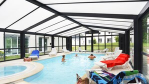 An indoor swimming pool with clear glass walls, surrounded by lounge chairs and a hot tub area.